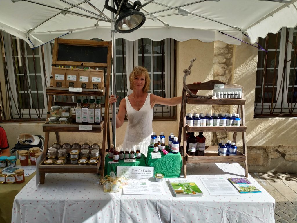 Contact Une artisane présente son stand de produits naturels au marché local. L’étal est organisé avec des huiles essentielles, tisanes bio, baumes artisanaux et confitures, sous un parasol blanc pour créer une ambiance conviviale et authentique.